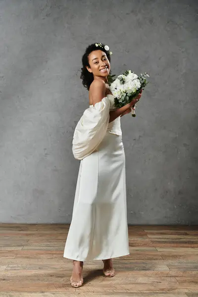 A beautiful African American bride in a white wedding dress with flowers in her hair, smiling and holding a bouquet. — Stock Photo