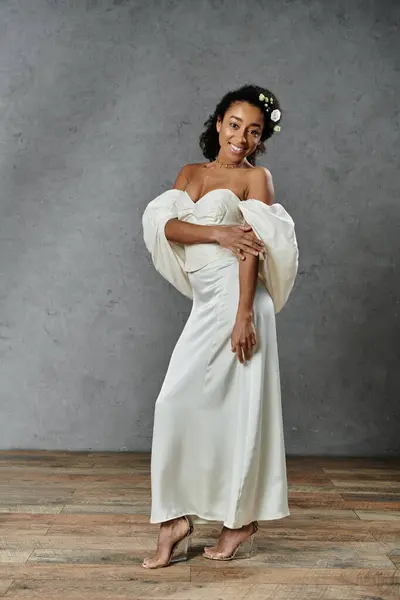 A beautiful African American bride poses in a flowing white wedding dress with flowers in her hair, against a grey backdrop. — Stock Photo