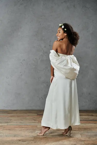 Una novia afroamericana con un vestido de novia blanco y una corona floral sonríe mientras está de pie sobre un fondo gris. - foto de stock