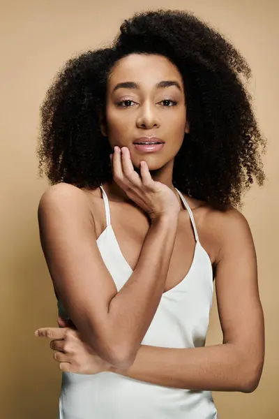A beautiful African American woman with natural makeup, wearing a white tank top, gazes softly at the camera against a beige background. — Stock Photo