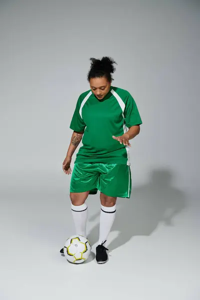 Una donna che indossa una maglia da calcio verde e pantaloncini gocciola un pallone da calcio su uno sfondo grigio. — Foto stock