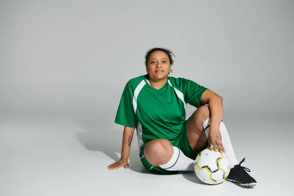 Una donna in jersey verde si siede con un pallone da calcio davanti a uno sfondo bianco. — Foto stock