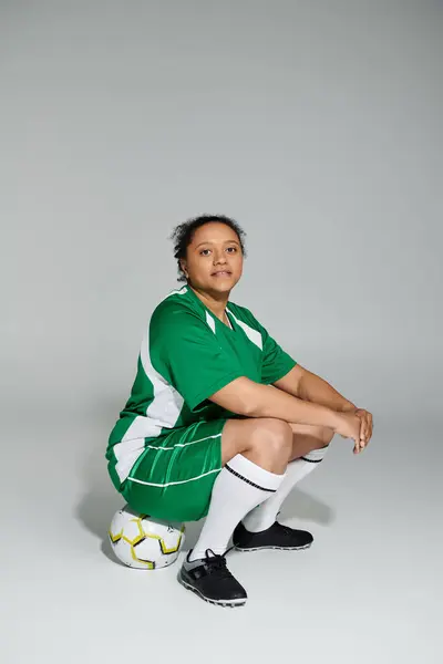 Una donna sicura di sé in uniforme da calcio verde siede pronta su un pallone da calcio in uno studio. — Foto stock