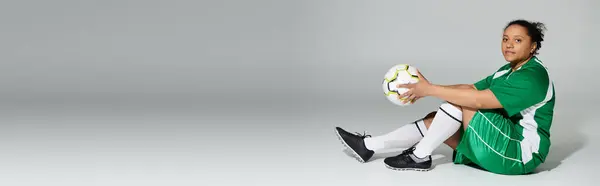 Una donna in uniforme verde si siede a terra, con un pallone da calcio in mano. — Foto stock