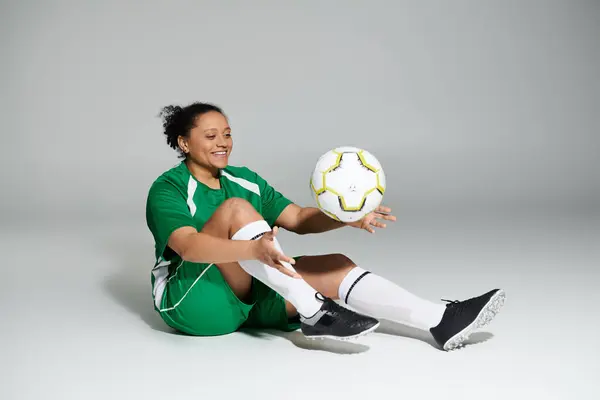 A woman in a green jersey sits on the ground, balancing a soccer ball on her leg. — Stock Photo