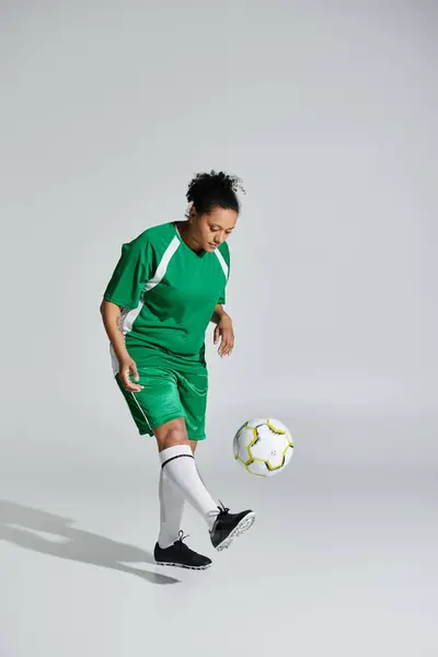 Un atleta donna in uniforme da calcio verde destreggia un pallone da calcio bianco in un ambiente da studio. — Foto stock