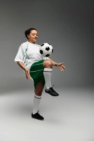 Una mujer en un jersey blanco y pantalones cortos verdes hace malabares con una pelota de fútbol en un ambiente de estudio. - foto de stock