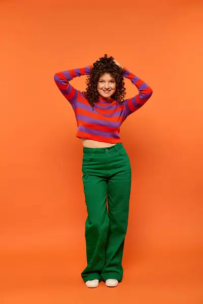 A young woman with curly hair, wearing a striped shirt and green pants, poses against a bright orange background. — Stock Photo