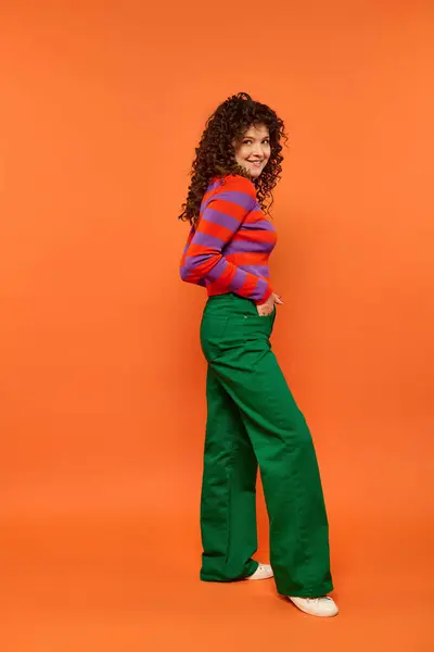 A young woman with curly hair poses in a striped sweater and green pants against a bright orange backdrop. — Stock Photo