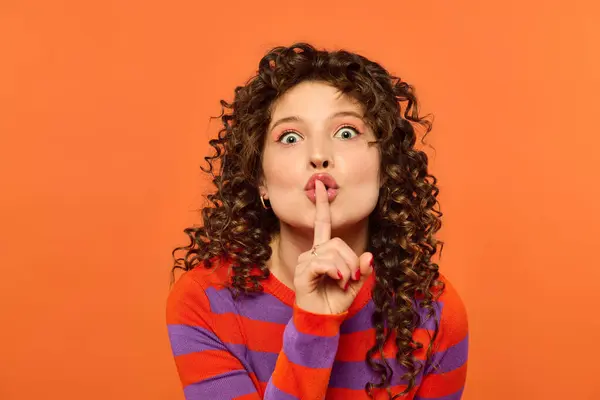 A young woman with curly hair and vibrant clothing holds her finger to her lips, whispering a secret against a bright orange background. — Stock Photo