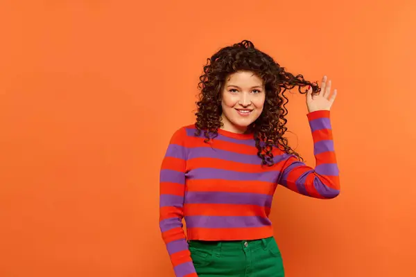 Curly-haired girl smiles against orange background in red and purple sweater and green pants. — Stock Photo