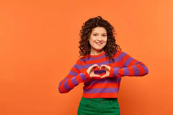 A young woman with curly hair poses against a bright orange background, forming a heart shape with her hands. — Stock Photo