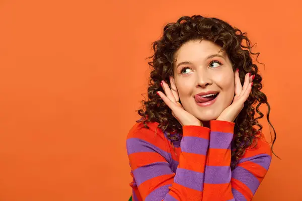 A young woman with curly hair poses in a striped sweater against a bright orange background, looking playful and expressive. — Stock Photo