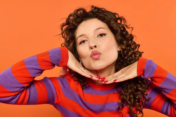 A young woman with curly hair poses against a bright orange background, wearing a striped sweater and pouting her lips. — Stock Photo