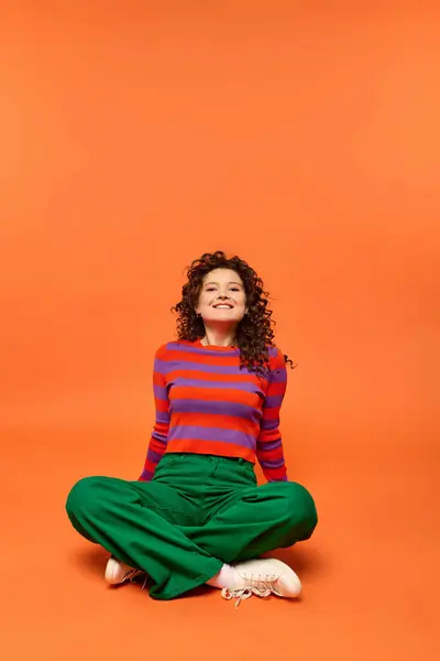 Young woman with curly hair, striped sweater and green pants, smiles and looks at camera with legs crossed in front of orange background. — Stock Photo