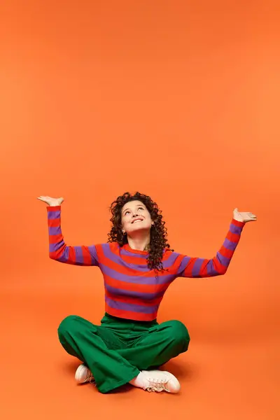 Curly-haired woman poses with arms raised sitting against a bright orange background, dressed in red and purple sweater with green pants. — Stock Photo