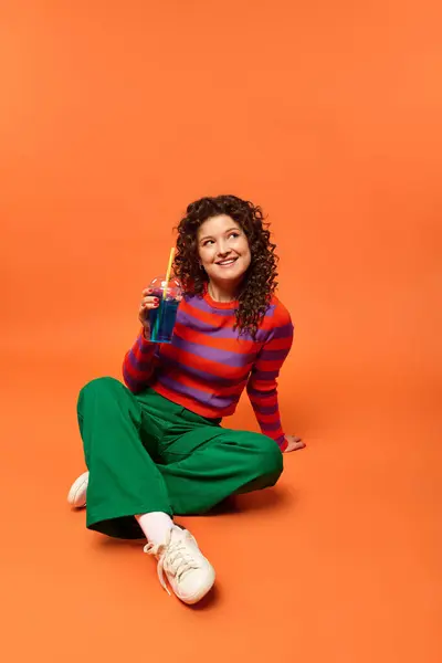 A young woman with curly hair sits with a blue drink, gazing up dreamily on a vibrant orange background. — Stock Photo