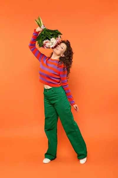 Una joven con el pelo rizado posa en un vibrante estudio naranja, sosteniendo un ramo de flores sobre su cabeza. - foto de stock