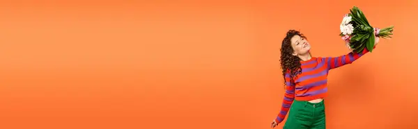 A young woman with curly hair smiles as she holds a bouquet of flowers in front of an orange background. — Stock Photo