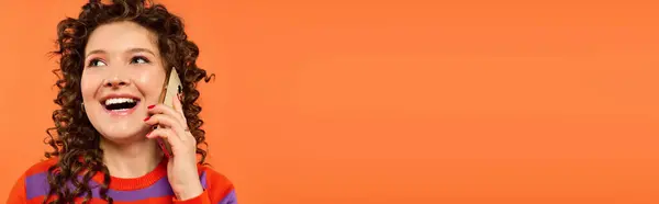 A young woman with curly hair is smiling while talking on her phone, on a bright orange background. — Stock Photo