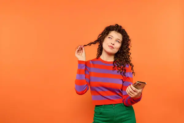 Une jeune femme aux cheveux bouclés pose sur un fond orange vif, tenant son téléphone et regardant doucement sur le côté. — Photo de stock