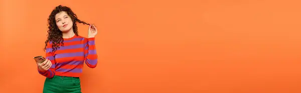 A young woman with curly hair poses against a bright orange background, holding a phone and twirling a lock of her hair. — Stock Photo
