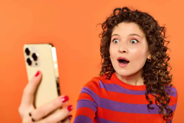 A shocked young woman with curly hair stares at her phone, wearing a striped sweater against an orange background. — Stock Photo