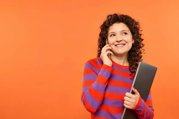 A young woman with curly hair smiles as she talks on her phone and holds a laptop against a vibrant orange background. — Stock Photo