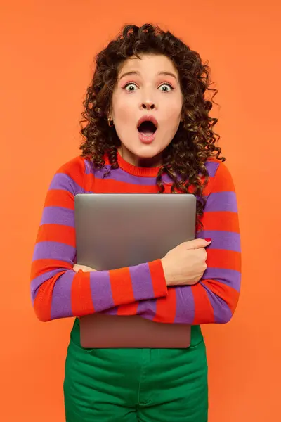 Curly-haired woman in a striped sweater and green pants stands against an orange background, holding a laptop with a surprised look. — Stock Photo