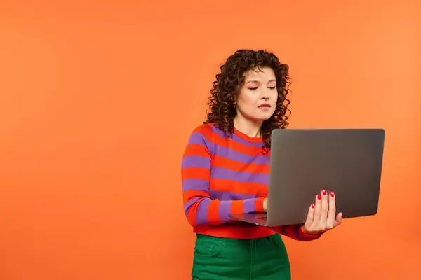 Mujer joven con el pelo rizado en suéter rojo y púrpura mira fijamente a la computadora portátil en el fondo naranja. - foto de stock