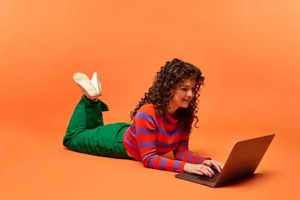 Una joven con el pelo rizado posa sobre un fondo naranja mientras usa un portátil. - foto de stock