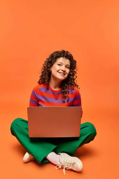 A young woman with curly hair sits with her legs crossed on a bright orange background, holding a laptop and smiling at the camera. — Stock Photo