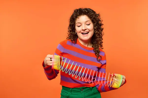 A young woman with curly hair is smiling and posing with a slinky toy in front of a bright orange background. — Stock Photo