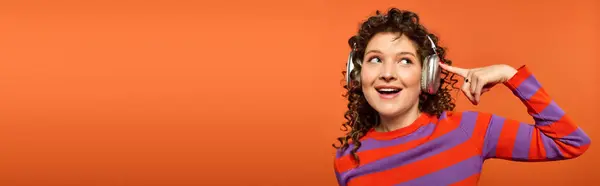 A young woman with curly hair and a vibrant sweater poses against a bright orange background, headphones on, enjoying the music. — Stock Photo