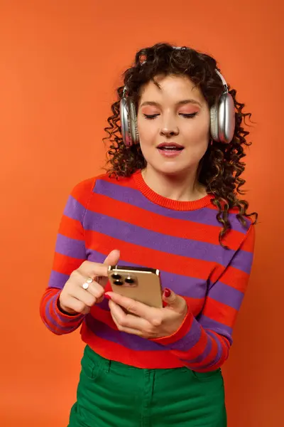 Mujer joven con ropa brillante y auriculares, con el pelo rizado, sonríe mientras usa el teléfono sobre fondo naranja. — Stock Photo