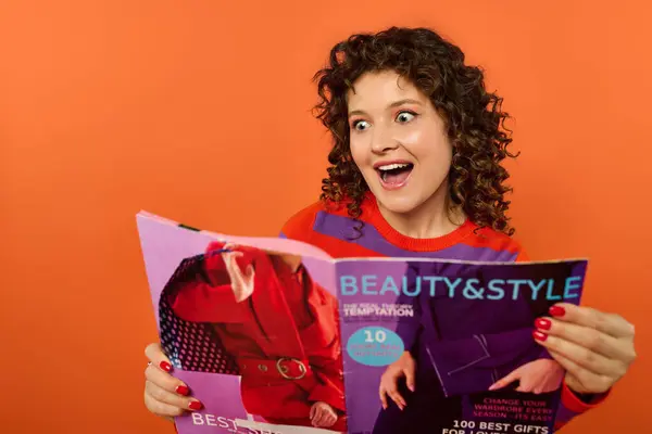 Young woman in her 20s poses excitedly in vibrant attire against orange background, reacting to magazine shes reading. — Stock Photo