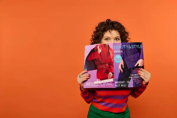 A young woman with curly hair poses in colorful attire against a bright orange background while reading a fashion magazine. — Stock Photo