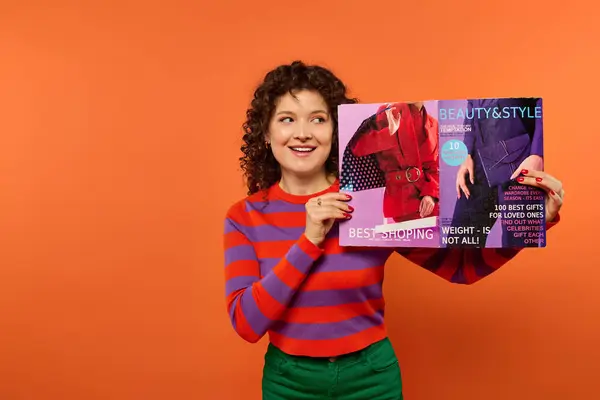 Lockige Frau hält Magazin vor orangefarbenem Hintergrund, trägt gestreiften Pullover und grüne Hose. — Stockfoto