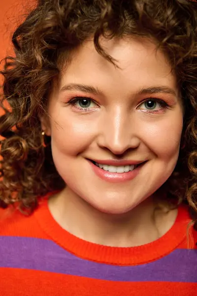 Retrato de close-up de uma jovem mulher com cabelo encaracolado, em suéter vermelho e roxo, sorrindo brilhantemente contra um fundo laranja brilhante. — Fotografia de Stock