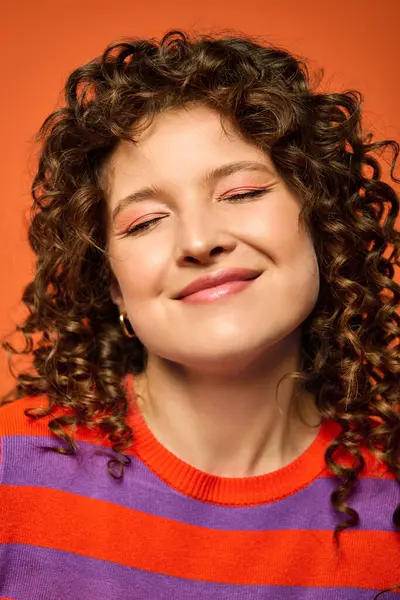 A young woman with curly brown hair smiles brightly while wearing a striped sweater against a vibrant orange background. — Stock Photo