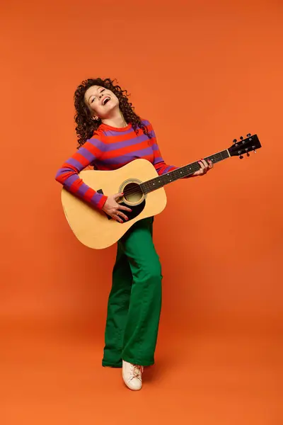 Une jeune femme aux cheveux bouclés joue de la guitare acoustique tout en souriant brillamment sur un fond orange vif. — Photo de stock
