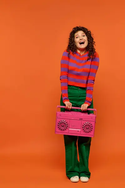 A woman with curly hair holds a pink boombox in front of an orange background, wearing a striped shirt, green pants, and white shoes. — Stock Photo