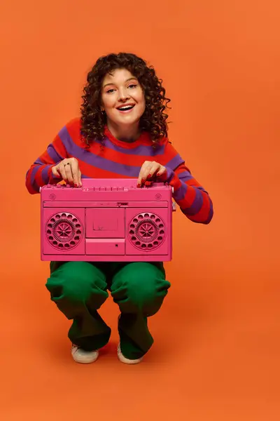 A young woman with curly hair poses playfully in front of an orange backdrop, holding a pink boombox. — Stock Photo
