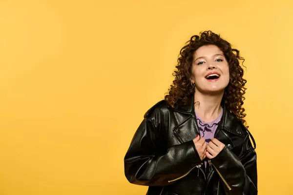 A young woman with curly hair poses in a black leather jacket against a vibrant yellow background. — Stock Photo
