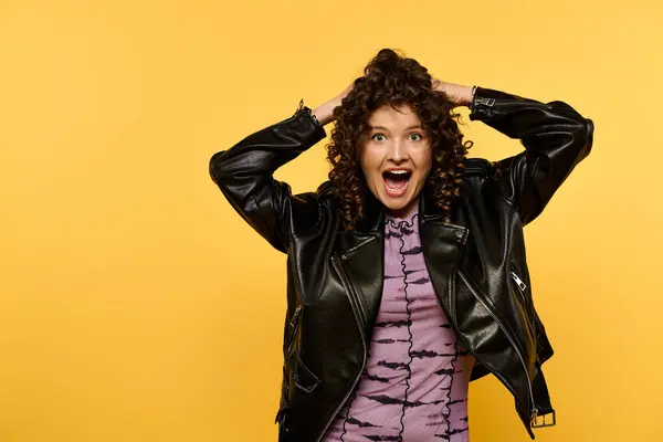 A young woman with curly hair and a black leather jacket poses against a yellow background, looking surprised and excited. — Stock Photo