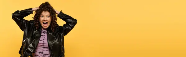 A young woman with curly hair wearing a black leather jacket stands against a bright yellow background, her mouth open in surprise. — Stock Photo