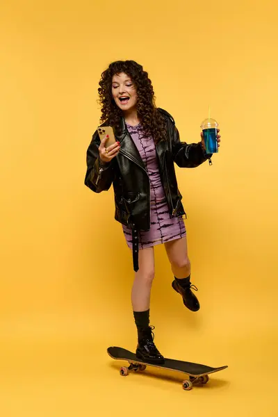 Une jeune femme élégante dans une veste en cuir pose en skateboard, tenant un smartphone et une boisson. — Photo de stock