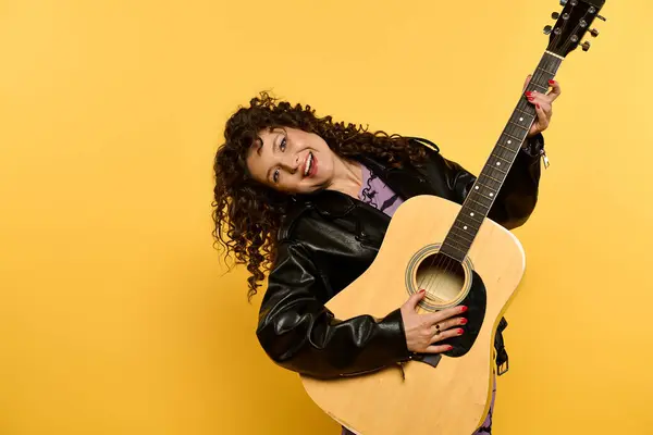 Une jeune femme élégante dans une veste en cuir pose avec une guitare sur un fond jaune vif. — Photo de stock