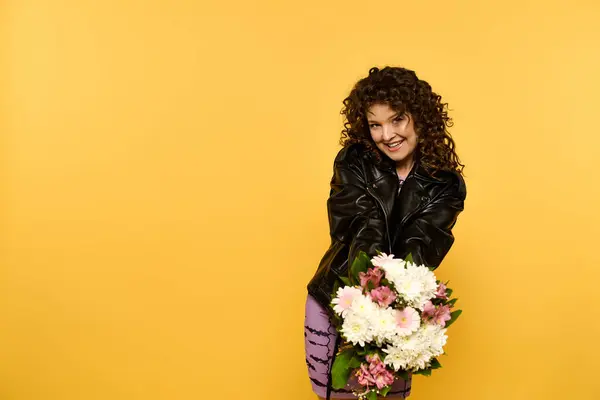 Uma jovem mulher com cabelos encaracolados posa em uma jaqueta de couro preto enquanto segura um buquê de flores contra um fundo amarelo brilhante. — Fotografia de Stock