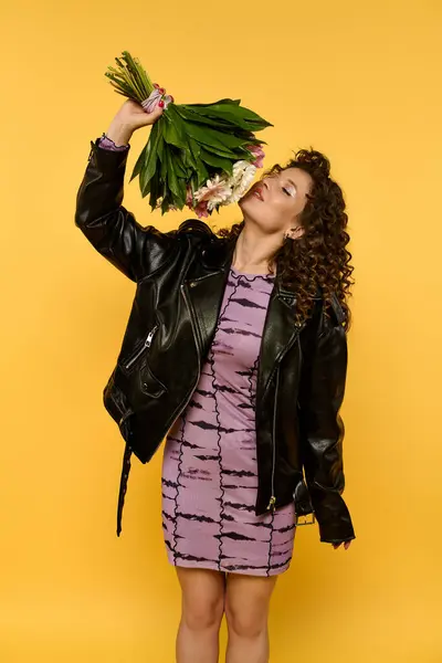 Una joven con una chaqueta de cuero negro y un vestido morado sostiene un ramo de flores y sonrisas mientras posa sobre un fondo amarillo. - foto de stock